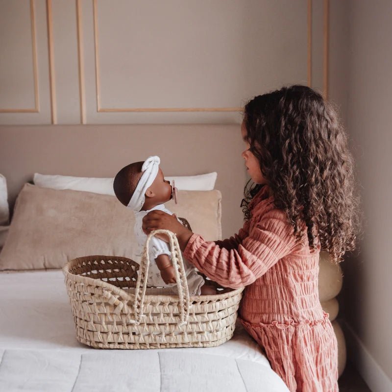 A girl with long curly dark hair sits on a bed beside a woven baby basket, holding a baby doll wearing a white outfit and headband in a softly lit bedroom.