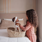 A girl with long curly dark hair sits on a bed beside a woven baby basket, holding a baby doll wearing a white outfit and headband in a softly lit bedroom.